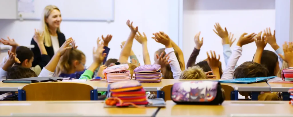 Kinder sitzen im Klassenzimmer und strecken ihre Arme in die Höhe. Im Vordergrund stehen Tische. Darauf liegen Federmäppchen.