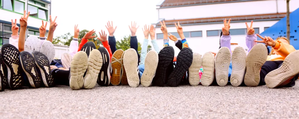 Kinder liegen nebeneinander auf dem Schulhof. Man sieht nur ihre Schusohlen und ihre Arme und Hände, die sie zur Begrüßung in die Höhe strecken.