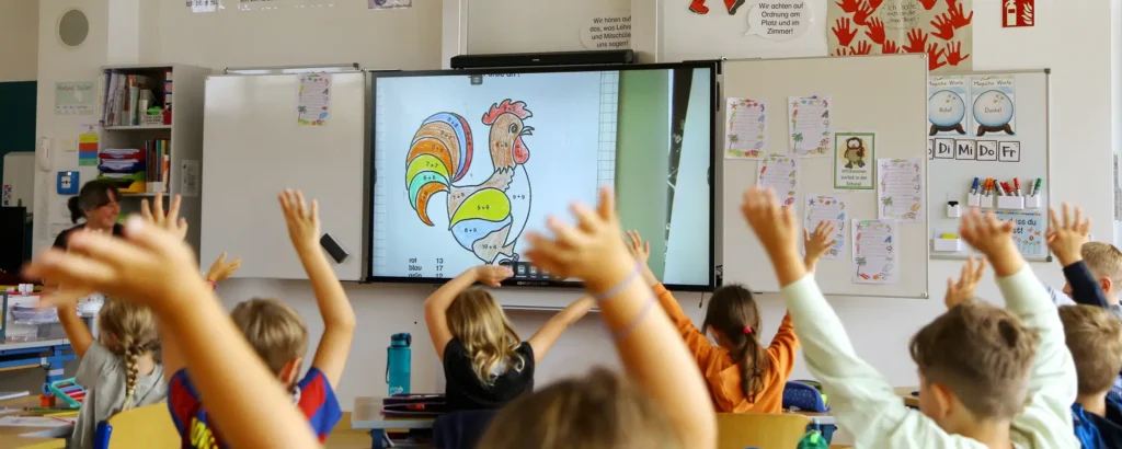 Schüler sitzen vor dem Whiteboard und strecken die Arme in die Höhe.