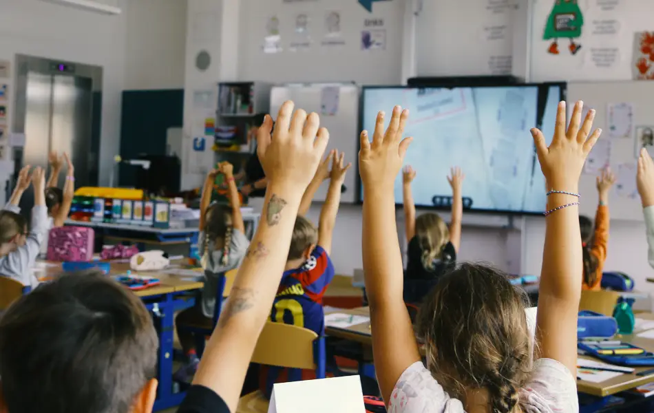 Kinder sitzen in ihrem Klassenzimmer und strecken die Arme in die Höhe.