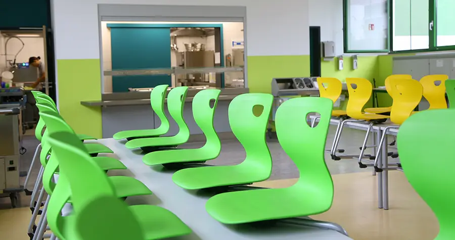 Die Mensa der Grundschule Zeitlarn. Im Hintergrund befindet sich die Essensausgabe. Im Vordergrund sind bunte Stühle, die auf die Tische gestellt sind.
