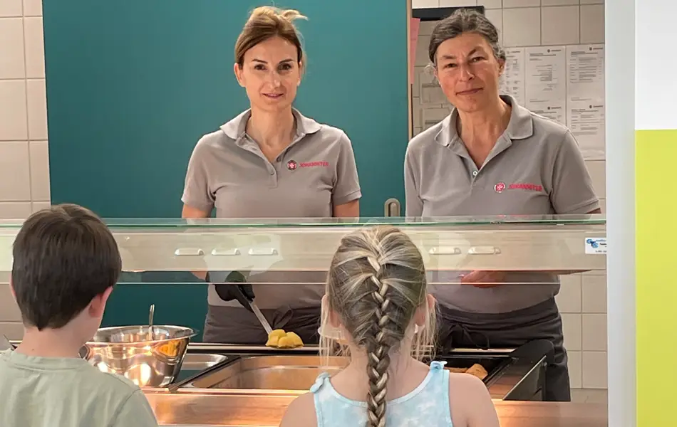 2 Mitglieder des Küchenteams und 2 Kinder an der Essensausgabe der Mensa.