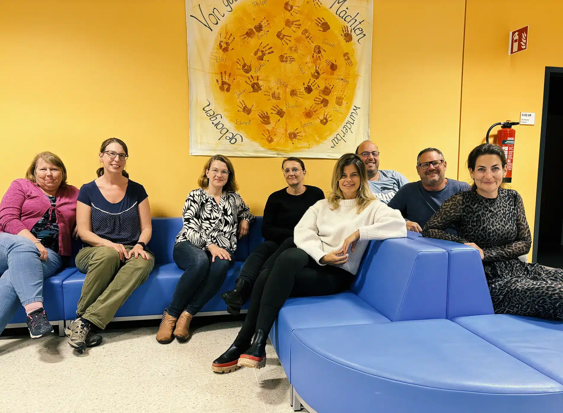 8 Mitglieder des Elternbeirats der Grundschule Zeitlarn sitzen auf den blauen Sofas im Schulflur.